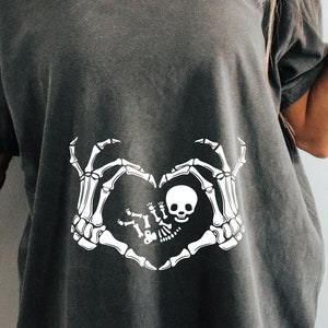 K&ouml;nnte beinhalten: Ein dunkelgraues T-Shirt mit einem wei&szlig;en Skelett-Hand-Design, das eine Herzform um ein Baby-Skelett bildet. Das Design enth&auml;lt den Text "Little Boo"