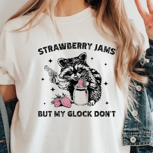 Pu&ograve; includere: Maglietta bianca con un'illustrazione in bianco e nero di un procione che tiene una pistola e un barattolo di marmellata di fragole. Il testo "Strawberry Jams But My Glock Don't" &egrave; stampato sulla maglietta.