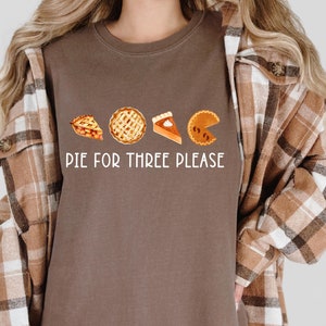 May include: Espresso-colored t-shirt featuring a watercolor design of four pie slices. The text "PIE FOR THREE PLEASE" is printed in white. The shirt is a crew neck style.