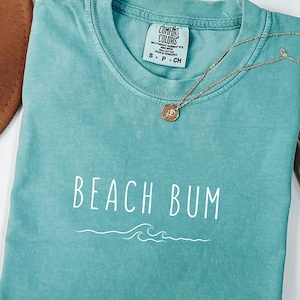 Może przedstawiać: Turkusowy t-shirt z napisem "Beach Bum" w białym kolorze i wzorem fali. Koszulka ma postarzony wygląd i jest wykonana z miękkiej, wygodnej tkaniny.
