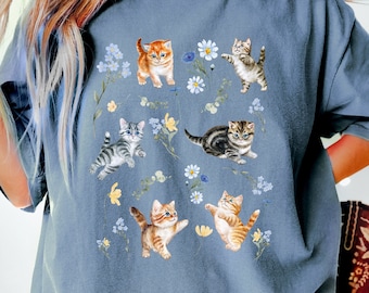 May include: A blue-grey t-shirt featuring a charming print of playful kittens amidst delicate flowers and foliage. The kittens are depicted in various poses, with orange, grey, and brown tabby patterns. The floral elements include white daisies and blue and yellow wildflowers.