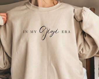 Op de afbeelding: Beige sweatshirt met de tekst "IN MY Gigi ERA" in zwart script. De sweatshirt heeft een ronde hals en lange mouwen. De afbeelding is een close-up van de sweatshirt.