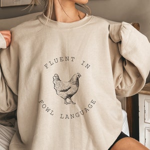 May include: A beige sweatshirt with a black graphic of a chicken and the text "Fluent in Fowl Language".
