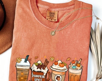Può includere: Una t-shirt color terracotta con una stampa grafica che raffigura quattro diverse bevande, tra cui una tazza con l'etichetta PUMPKIN SPICE, circondata da elementi autunnali come zucche, foglie e ghiande. La t-shirt presenta un girocollo.