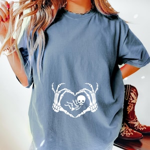 K&ouml;nnte beinhalten: Ein hellblaues T-Shirt mit einem wei&szlig;en Skelett-Hand-Design, das eine Herzform um ein Baby-Skelett bildet. Das Design enth&auml;lt den Text "Little Bones".