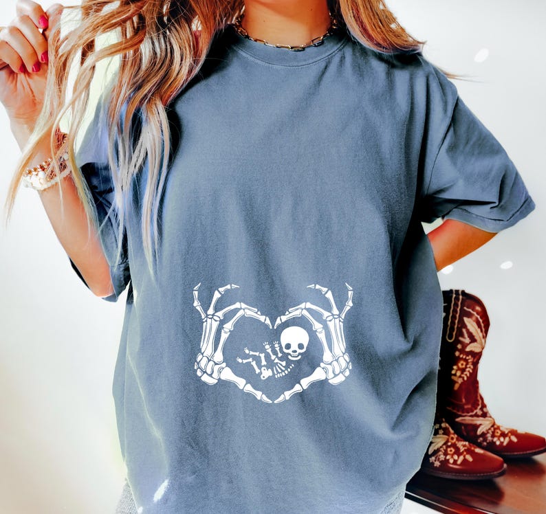 K&ouml;nnte beinhalten: Ein hellblaues T-Shirt mit einem wei&szlig;en Skelett-Hand-Design, das eine Herzform um ein Baby-Skelett bildet. Das Design enth&auml;lt den Text "Little Bones".