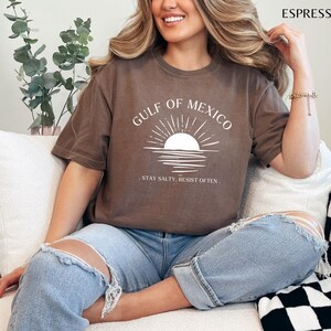 May include: A brown t-shirt with a white graphic of a sunset over water and the text "GULF OF MEXICO. STAY SALTY. RESIST OFTEN."