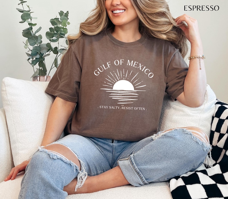 May include: A brown t-shirt with a white graphic of a sunset over water and the text "GULF OF MEXICO. STAY SALTY. RESIST OFTEN."