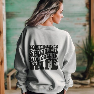 May include: A gray sweatshirt with the text "Somebody's Spoiled Blue Collar Wife" in black retro-style lettering.