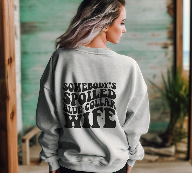 May include: A gray sweatshirt with the text "Somebody's Spoiled Blue Collar Wife" in black retro-style lettering.