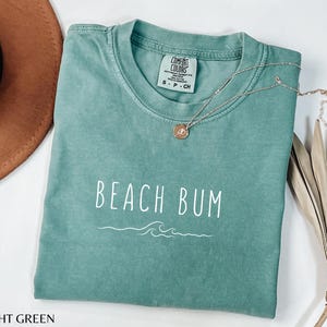 Może przedstawiać: Jasnozielony T-shirt z napisem "Beach Bum" w białym kolorze i wzorem fali. Koszulka wykonana jest z miękkiego, wygodnego materiału.
