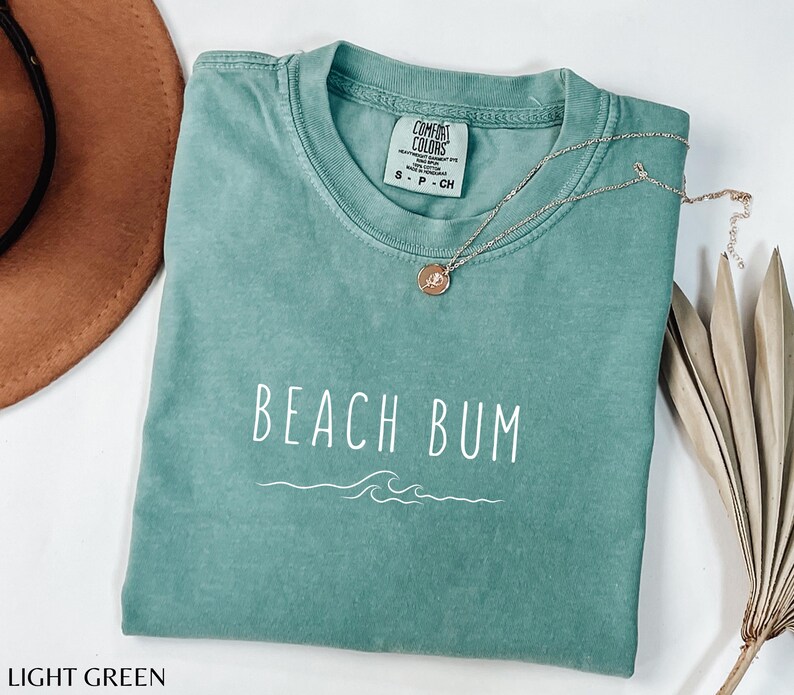 Może przedstawiać: Jasnozielony T-shirt z napisem "Beach Bum" w białym kolorze i wzorem fali. Koszulka wykonana jest z miękkiego, wygodnego materiału.