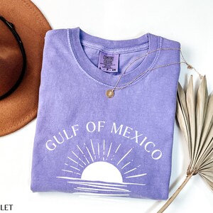 May include: A purple t-shirt with the text "Gulf of Mexico" in white lettering. The shirt features a white silhouette of a sunset with rays of light.