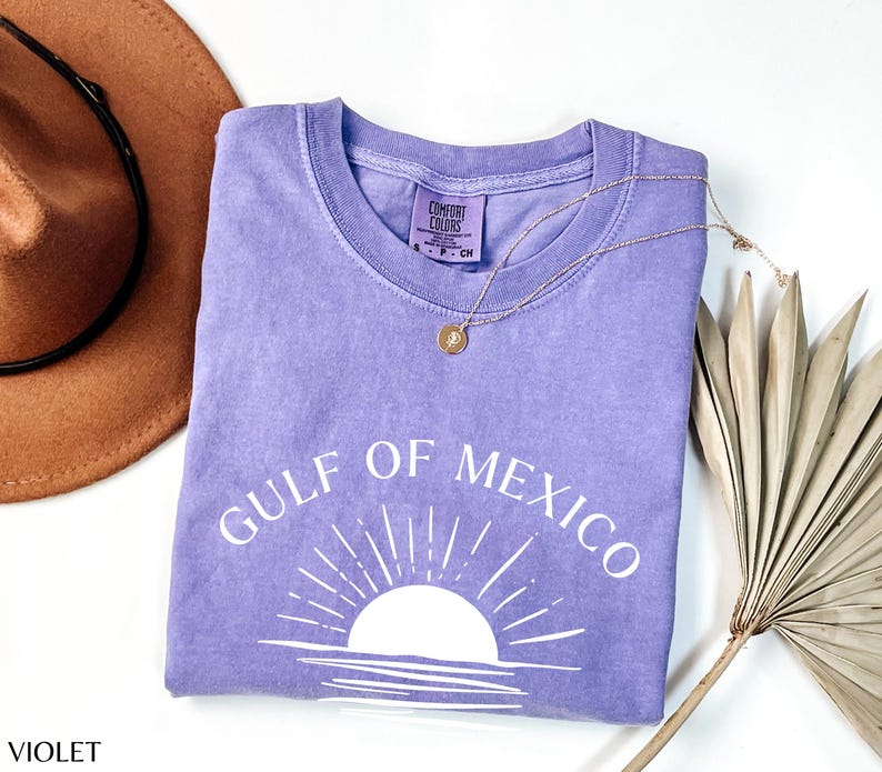 May include: A purple t-shirt with the text "Gulf of Mexico" in white lettering. The shirt features a white silhouette of a sunset with rays of light.