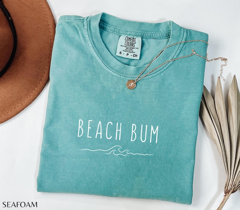 Może przedstawiać: Turkusowy t-shirt z napisem "Beach Bum" w białym kolorze i wzorem fali. Koszulka ma postarzony wygląd i jest wykonana z miękkiej, wygodnej tkaniny.