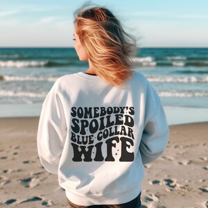 May include: White sweatshirt with black text that reads "Somebody's Spoiled Blue Collar Wife" in a retro font.
