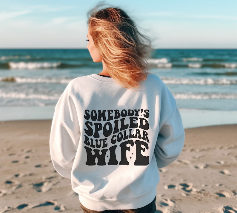 May include: White sweatshirt with black text that reads "Somebody's Spoiled Blue Collar Wife" in a retro font.