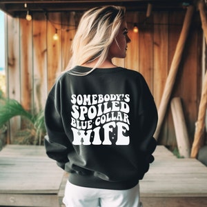 May include: Black sweatshirt with white text that reads "Somebody's Spoiled Blue Collar Wife" in a retro font.