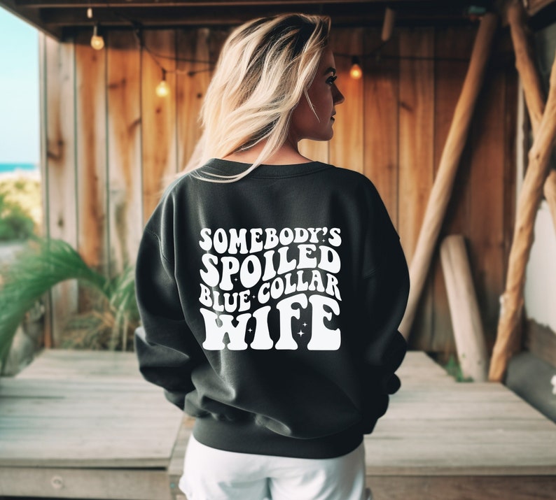 May include: Black sweatshirt with white text that reads "Somebody's Spoiled Blue Collar Wife" in a retro font.