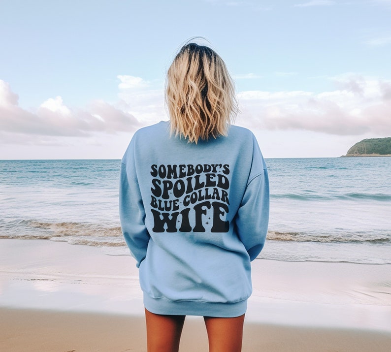 May include: A light blue sweatshirt with the text "Somebody's Spoiled Blue Collar Wife" in black.