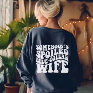 May include: A dark blue sweatshirt with white text that reads "Somebody's Spoiled Blue Collar Wife" in a retro font.