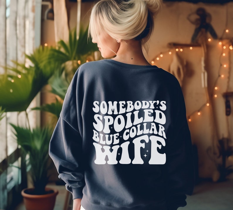 May include: A dark blue sweatshirt with white text that reads "Somebody's Spoiled Blue Collar Wife" in a retro font.