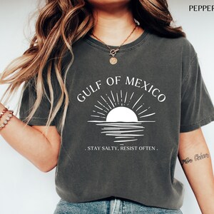 May include: A dark gray t-shirt with a white graphic of a sunset over the Gulf of Mexico. The text "Gulf of Mexico" is written in a white arc above the graphic. The text ". Stay Salty. Resist Often." is written below the graphic.