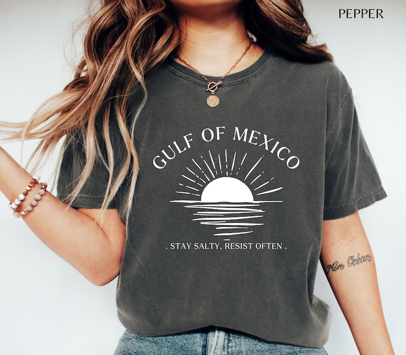 May include: A dark gray t-shirt with a white graphic of a sunset over the Gulf of Mexico. The text "Gulf of Mexico" is written in a white arc above the graphic. The text ". Stay Salty. Resist Often." is written below the graphic.