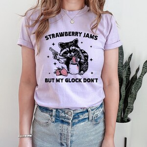 Pu&ograve; includere: Una maglietta color lavanda con una grafica in bianco e nero di un procione che tiene una pistola e un barattolo di marmellata di fragole. Il testo "Strawberry Jams But My Glock Don't" &egrave; stampato sulla maglietta.