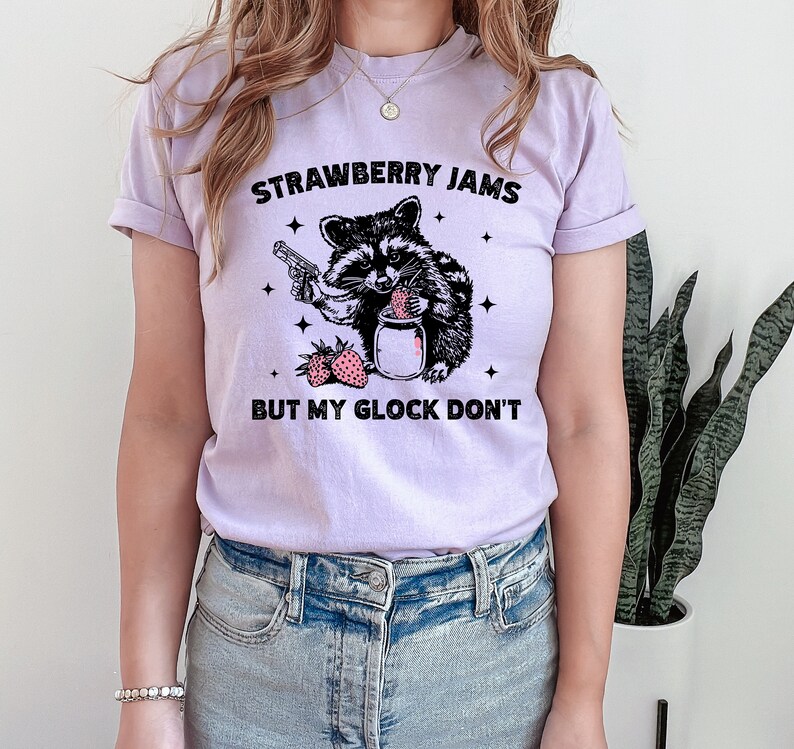 Pu&ograve; includere: Una maglietta color lavanda con una grafica in bianco e nero di un procione che tiene una pistola e un barattolo di marmellata di fragole. Il testo "Strawberry Jams But My Glock Don't" &egrave; stampato sulla maglietta.