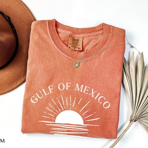 May include: A coral-colored t-shirt with a white graphic of a sunset over the Gulf of Mexico. The text "GULF OF MEXICO" is written in white above the graphic.