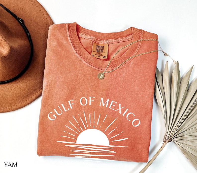 May include: A coral-colored t-shirt with a white graphic of a sunset over the Gulf of Mexico. The text "GULF OF MEXICO" is written in white above the graphic.