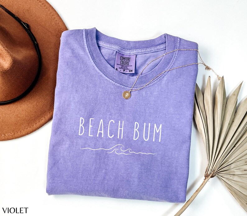 Może przedstawiać: Fioletowy T-shirt z napisem "Beach Bum" w białym kolorze i wzorem fali. Koszulka wykonana jest z miękkiego, wygodnego materiału.