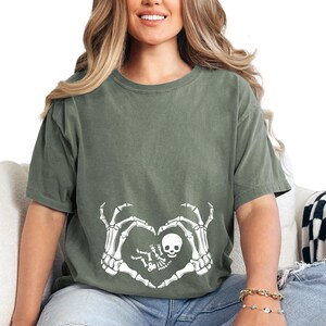 K&ouml;nnte beinhalten: Ein gr&uuml;nes T-Shirt mit einem wei&szlig;en Skelett-Hand-Design, das eine Herzform um ein Skelett-Baby bildet. Das Design ist mittig auf dem T-Shirt platziert.