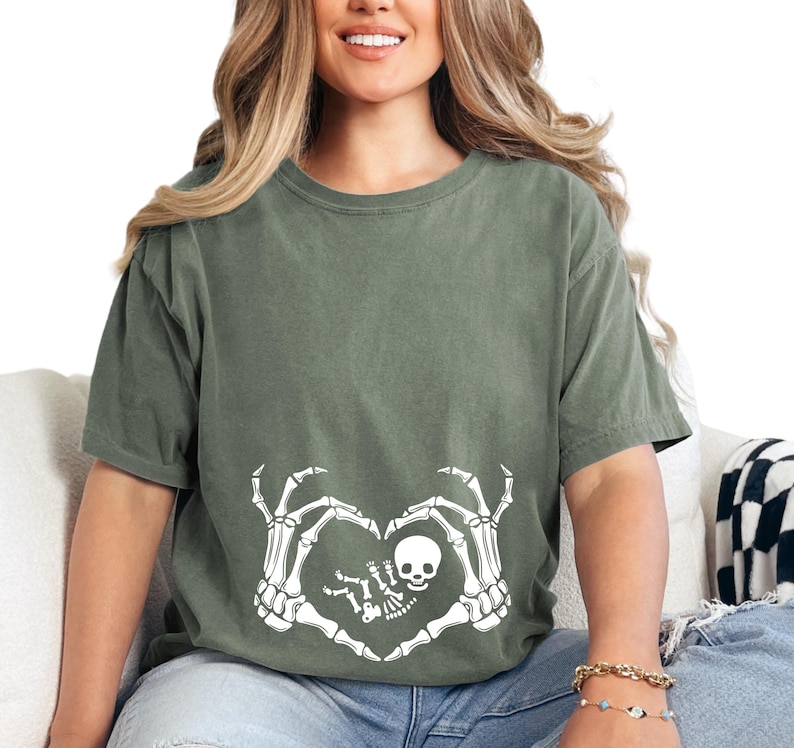 K&ouml;nnte beinhalten: Ein gr&uuml;nes T-Shirt mit einem wei&szlig;en Skelett-Hand-Design, das eine Herzform um ein Skelett-Baby bildet. Das Design ist mittig auf dem T-Shirt platziert.