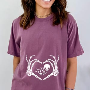 K&ouml;nnte beinhalten: Ein lila T-Shirt mit einem wei&szlig;en Skelett-Hand-Design. Die Skeletth&auml;nde bilden eine Herzform um ein kleines Skelett-Baby.