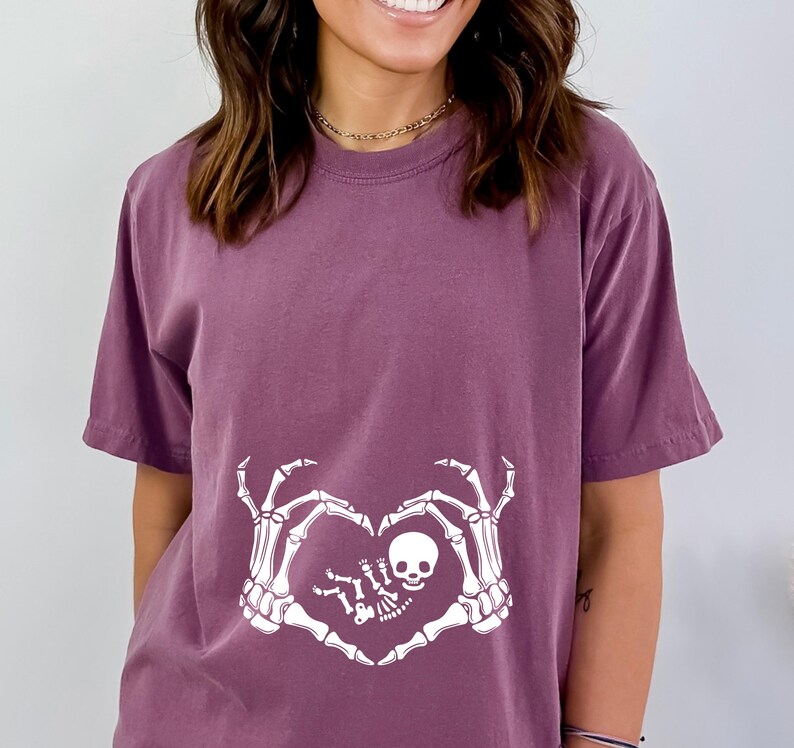 K&ouml;nnte beinhalten: Ein lila T-Shirt mit einem wei&szlig;en Skelett-Hand-Design. Die Skeletth&auml;nde bilden eine Herzform um ein kleines Skelett-Baby.