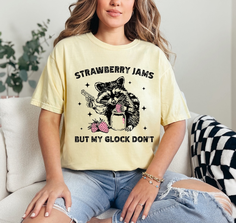 Pu&ograve; includere: Una maglietta gialla con un'illustrazione in bianco e nero di un procione che tiene una pistola e un barattolo di marmellata di fragole. Il testo "Strawberry Jams But My Glock Don't" &egrave; stampato sulla maglietta.