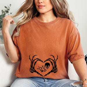 K&ouml;nnte beinhalten: Ein orangefarbenes T-Shirt mit einem schwarzen Skelett-Hand-Herz-Design. Die Skeletth&auml;nde halten ein kleines Skelett-Baby mit einem Sch&auml;delkopf.
