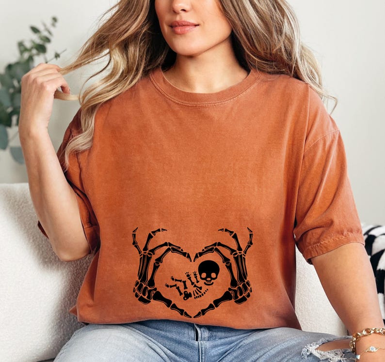 K&ouml;nnte beinhalten: Ein orangefarbenes T-Shirt mit einem schwarzen Skelett-Hand-Herz-Design. Die Skeletth&auml;nde halten ein kleines Skelett-Baby mit einem Sch&auml;delkopf.