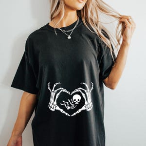 K&ouml;nnte beinhalten: Schwarzes T-Shirt mit einem wei&szlig;en Skelett-Hand-Design, das eine Herzform um ein Baby-Skelett bildet. Das Design sagt "Little Boo"