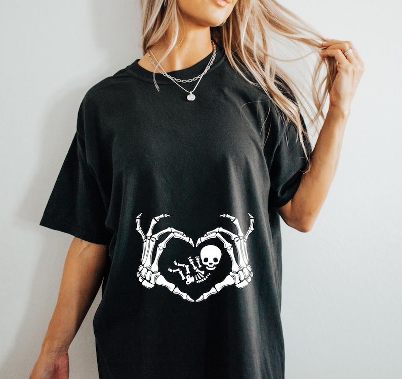 K&ouml;nnte beinhalten: Schwarzes T-Shirt mit einem wei&szlig;en Skelett-Hand-Design, das eine Herzform um ein Baby-Skelett bildet. Das Design sagt "Little Boo"