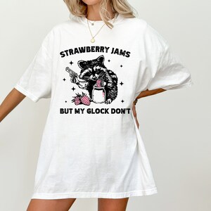 Pu&ograve; includere: Maglietta bianca oversize con una grafica nera di un procione che tiene un barattolo di marmellata di fragole e una pistola. Il testo recita "Strawberry Jams But My Glock Don't".