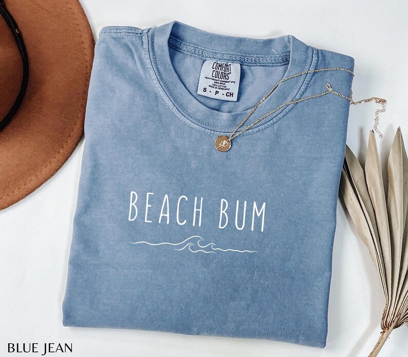 Może przedstawiać: Jasnoniebieska koszulka z napisem "Beach Bum" w białym kolorze i wzorem fal. Koszulka jest marki Comfort Colors i dostępna w rozmiarach S-P-CH.