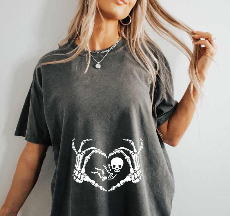 K&ouml;nnte beinhalten: Ein dunkelgraues T-Shirt mit einem wei&szlig;en Skelett-Hand-Design, das eine Herzform um ein Baby-Skelett bildet. Das Design enth&auml;lt den Text "Little Boo"