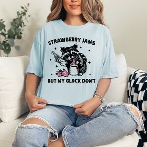Pu&ograve; includere: Una maglietta blu chiaro con un'illustrazione in bianco e nero di un procione che tiene una pistola e un barattolo di marmellata di fragole. Il testo "Strawberry Jams But My Glock Don't" &egrave; stampato sulla maglietta.