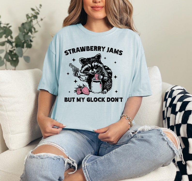 Pu&ograve; includere: Una maglietta blu chiaro con un'illustrazione in bianco e nero di un procione che tiene una pistola e un barattolo di marmellata di fragole. Il testo "Strawberry Jams But My Glock Don't" &egrave; stampato sulla maglietta.