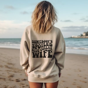 May include: A beige sweatshirt with the text "Somebody's Spoiled Blue Collar Wife" printed on the back in black.