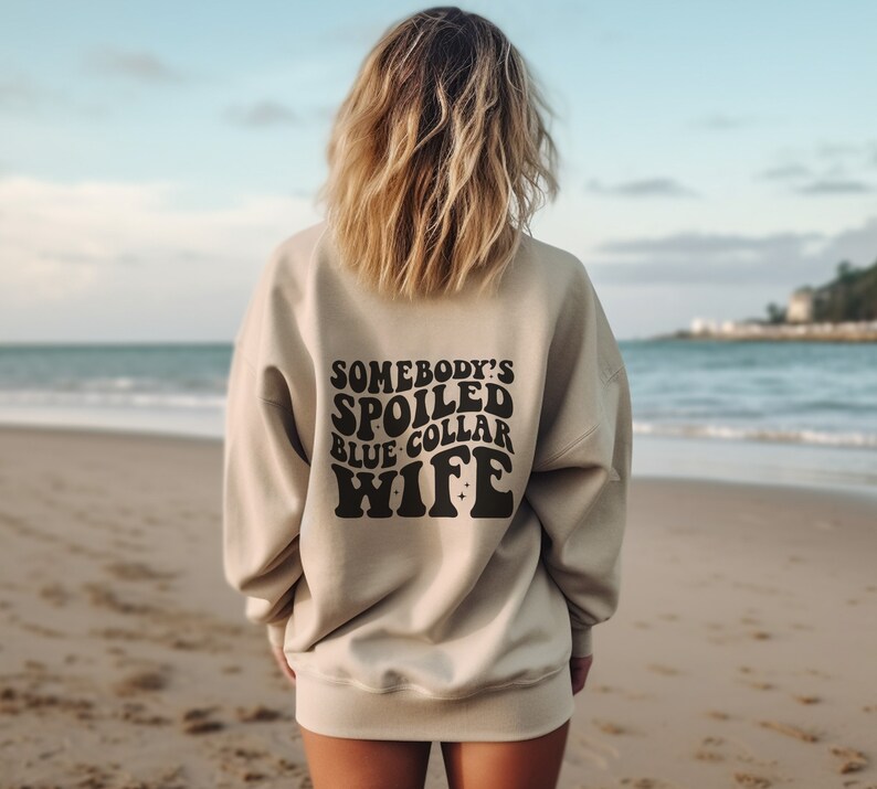 May include: A beige sweatshirt with the text "Somebody's Spoiled Blue Collar Wife" printed on the back in black.