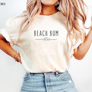 Może przedstawiać: Biały t-shirt z nadrukiem "Beach Bum" i małym wzorem fali z przodu.